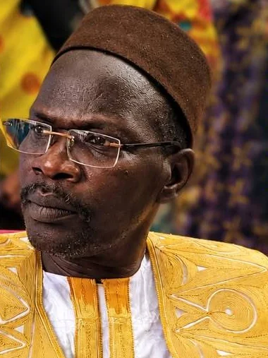 Portrait of a man wearing rimless glasses, a brown cap and a white boubou with gold embroidery