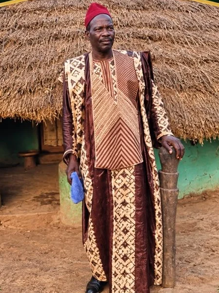 Marabout Sylla in an embroidered brown-and-cream boubou and a red cap, standing in front of a round thatched hut