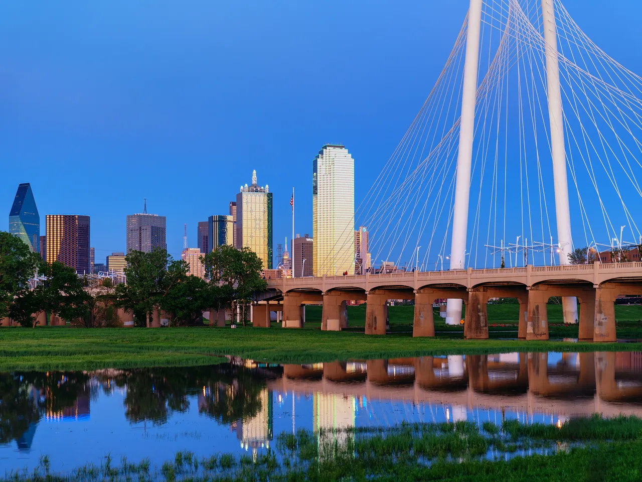 The Dallas skyline at dusk, reflected in the Trinity River beside the white arch of the Margaret Hunt Hill Bridge