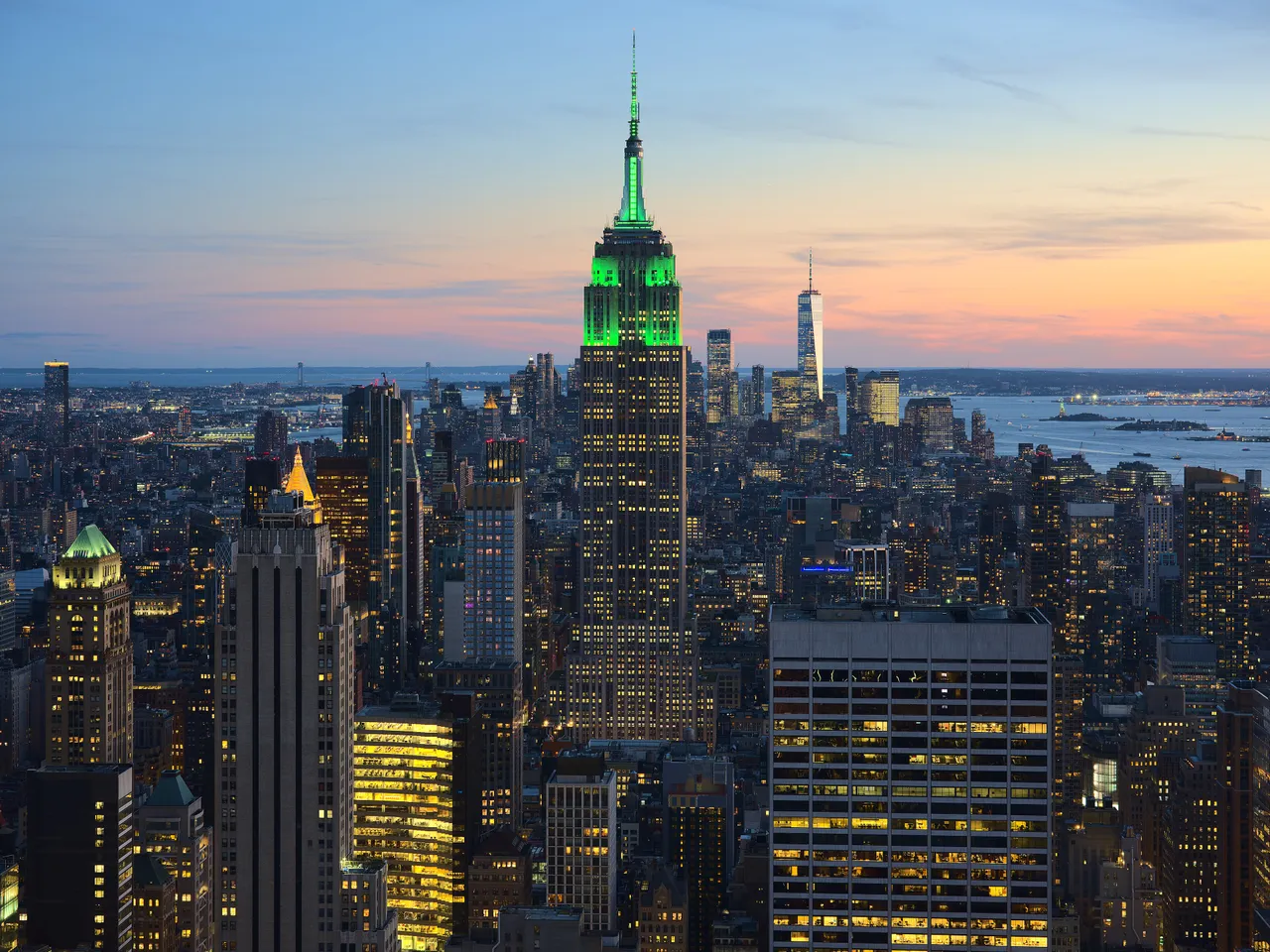 The Empire State Building lit in green at dusk above Midtown Manhattan, with One World Trade Center and the harbor in the distance